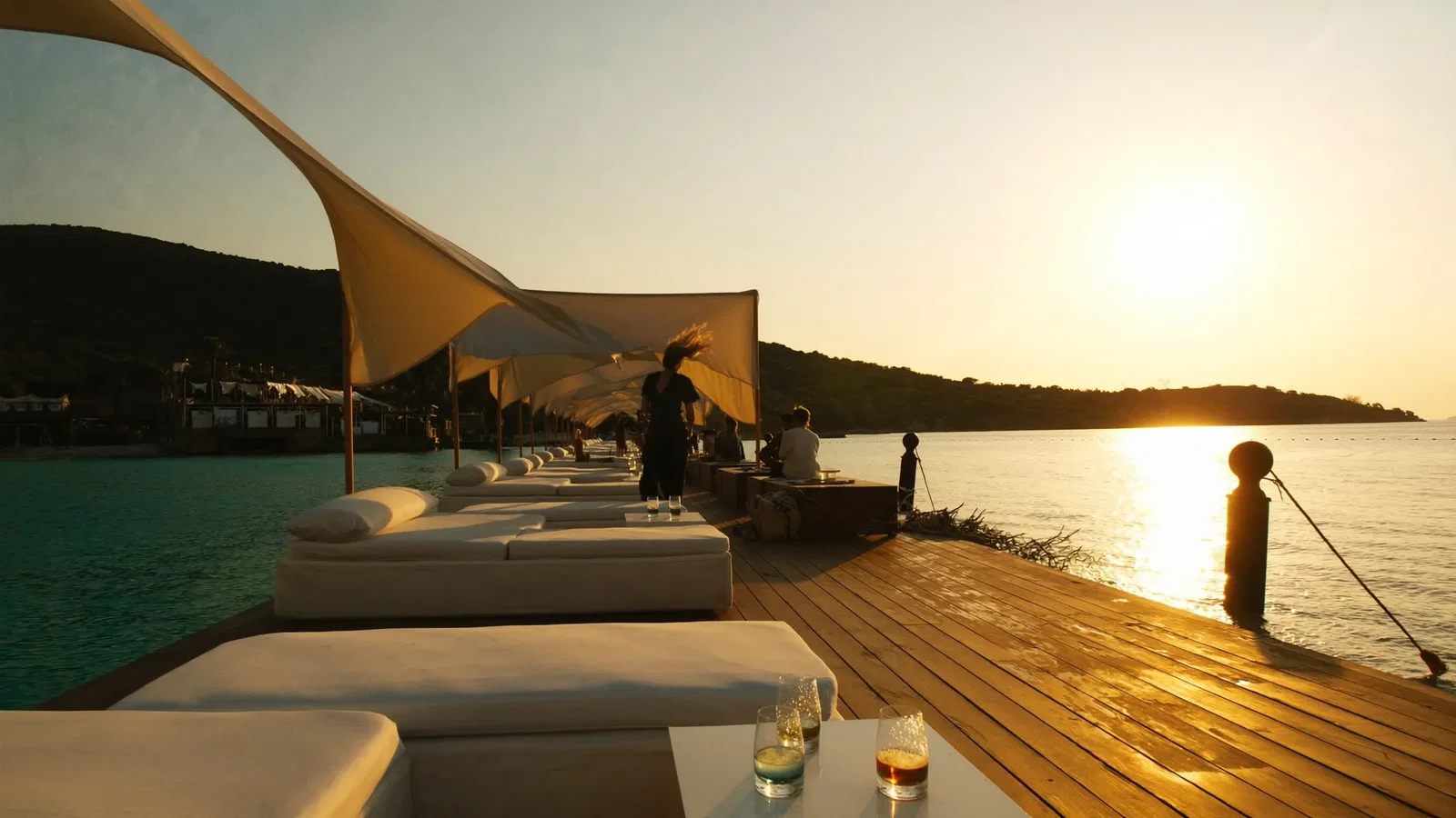 Daybeds and linen canopies on a wooden deck over turquoise water at a Bodrum beach club in golden-hour light