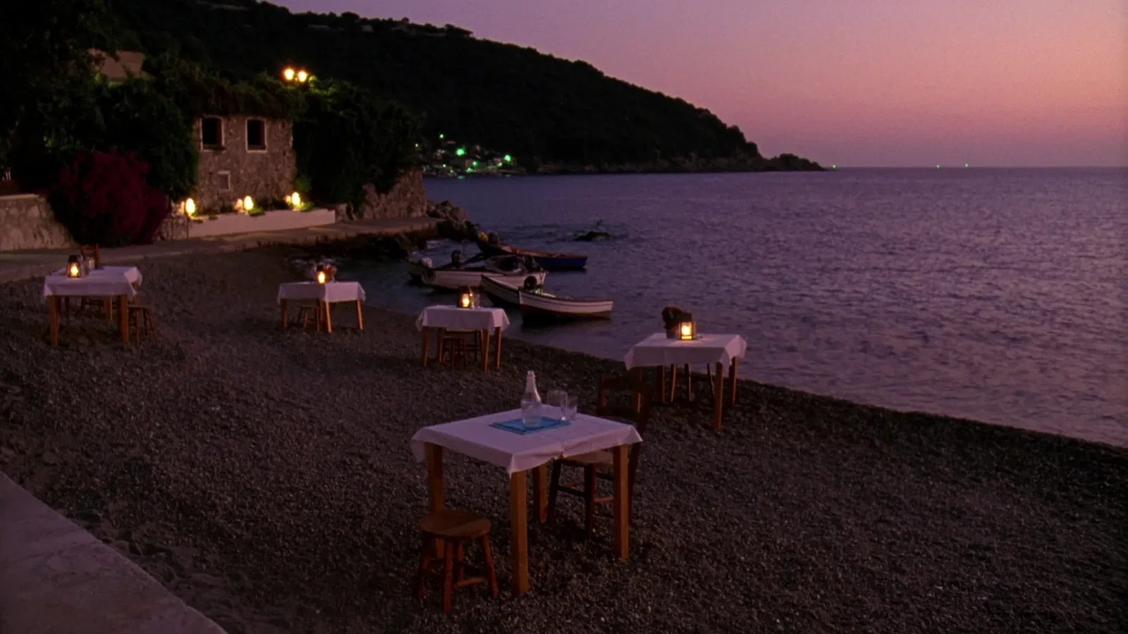 Simple tables with white linen and lanterns set out on the shingle at the water's edge in an Aegean fishing village at dusk