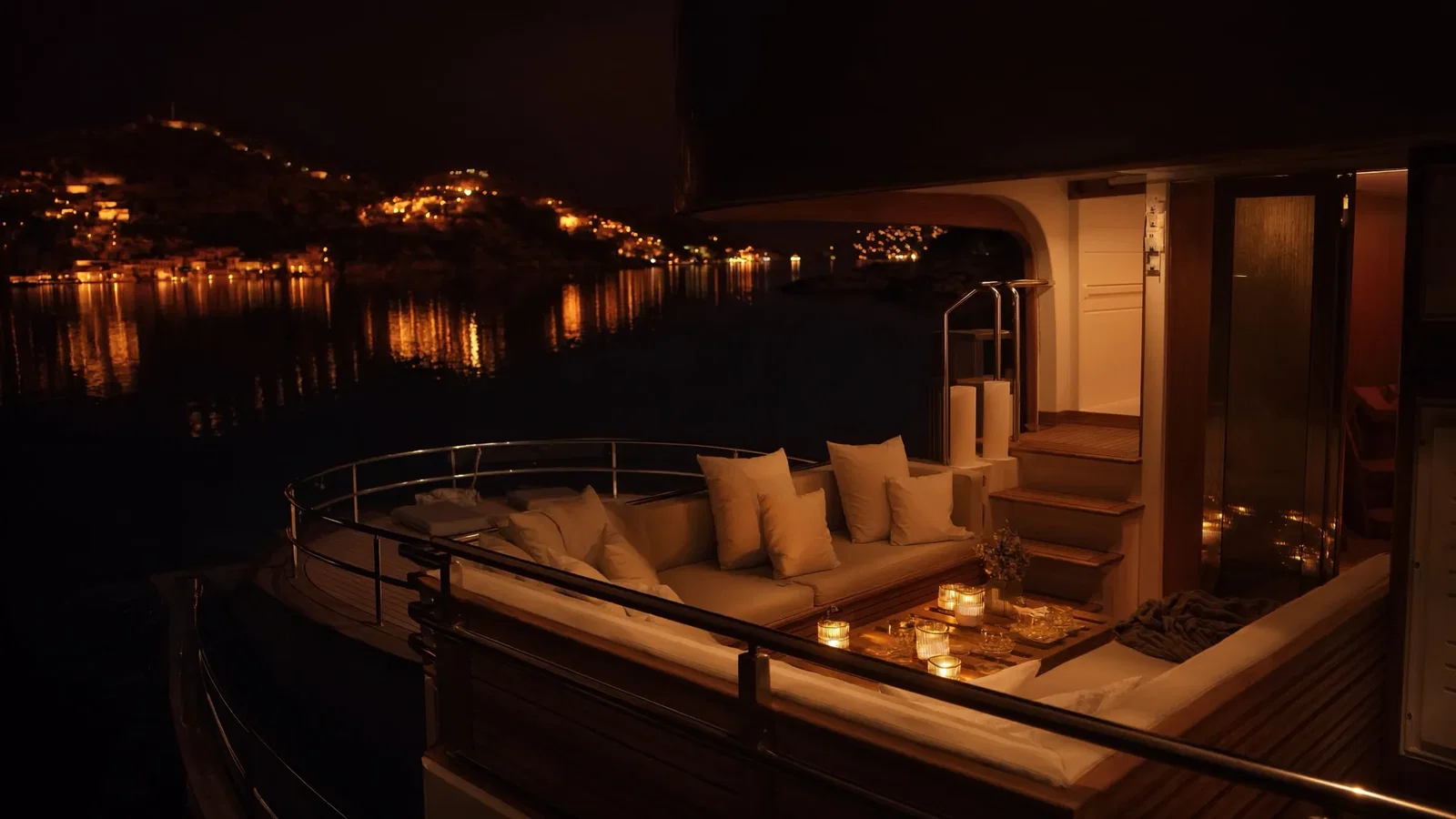 The lantern-lit aft deck of a yacht at night with cushions and the lights of a hillside town across the water