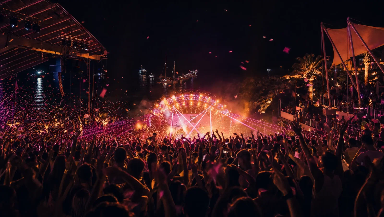 A packed open-air seaside nightclub after midnight with lasers, confetti and yachts anchored beyond the stage